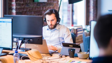 En man sittandes i kontorsmiljö med dator och headset.